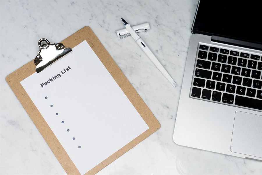 A clipboard with a blank packing list next to a laptop and pen on a marble desk